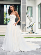 A woman modeling a strapless white wedding gown with a fitted bodice, lace-up front, and full flowing skirt with a long train, standing outdoors in front of a modern building facade. / Luxe matte satin has been used to create this beautiful A-line ballgown. The bodice features a plunging center front neckline that has been adorned with pearl encrusted bows. The full skirt is semi-circle cut and has pockets at the side seam.