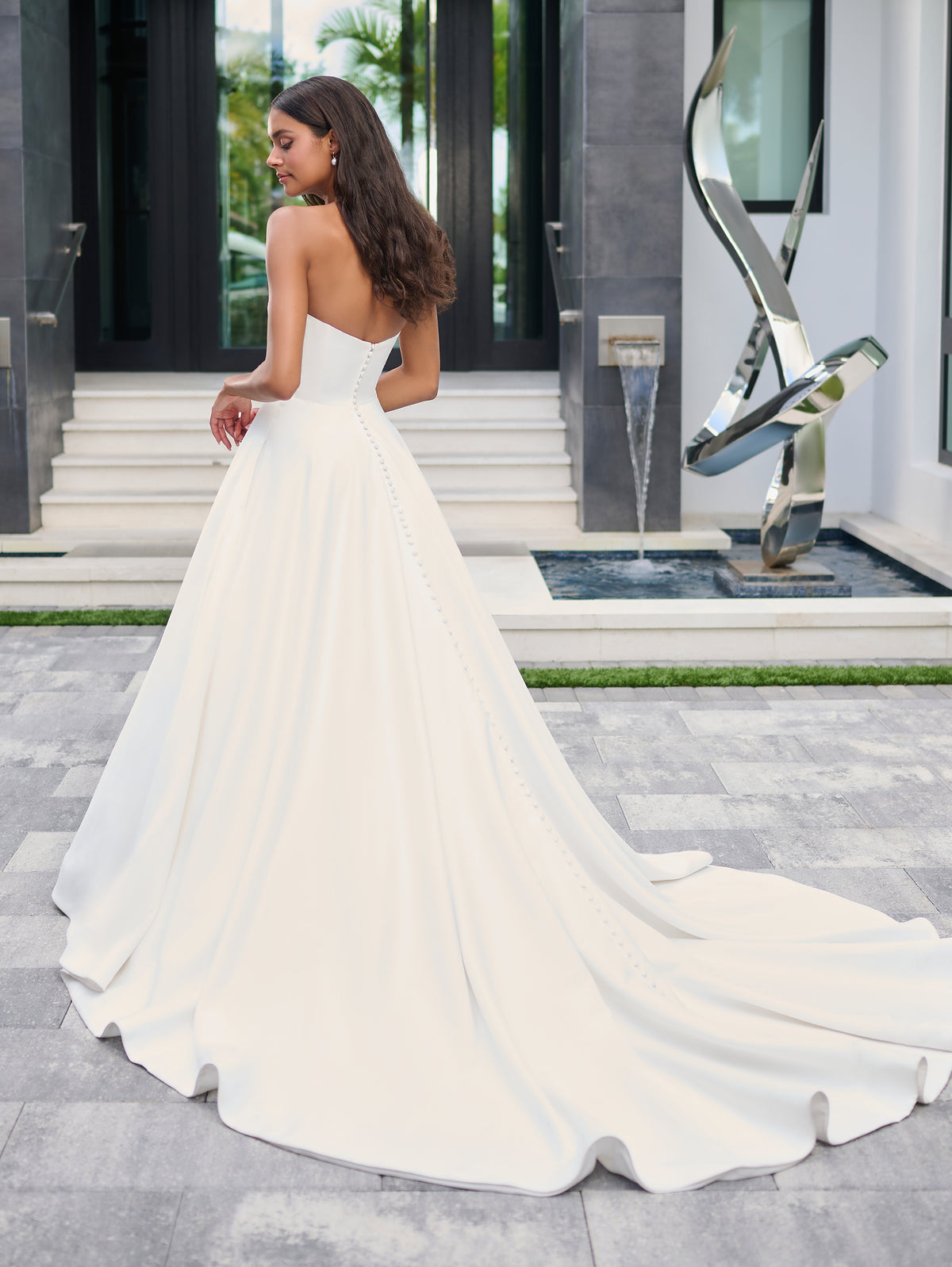 A woman wearing a strapless white ball gown with a long train, photographed outdoors in front of a modern building entrance with steps and a sculptural fountain. / Luxe matte satin has been used to create this beautiful A-line ballgown. The bodice features a plunging center front neckline that has been adorned with pearl encrusted bows. The full skirt is semi-circle cut and has pockets at the side seam.