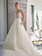 A woman modeling a sleeveless white wedding gown with a deep V-neckline, lace-embellished bodice, and a full flowing skirt with pockets, standing in a modern interior with a large window and sculptural decor. / Hand beaded bodice made of strings of various sized pearls with beaded edge finishing. There are sheer inserts at the side and a full illusion back bodice. The skirt is made a matte organza with flat pleats to create soft volume. A soft, pleated organza belt wraps around the waist