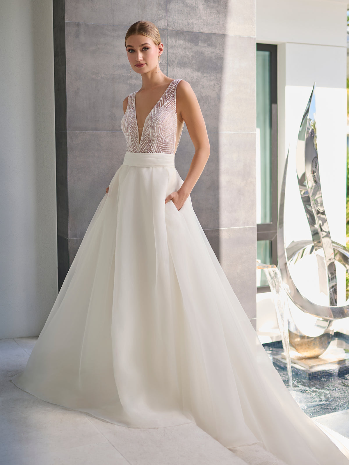 A woman modeling a sleeveless white wedding gown with a deep V-neckline, lace-embellished bodice, and a full flowing skirt with pockets, standing in a modern interior with a large window and sculptural decor. / Hand beaded bodice made of strings of various sized pearls with beaded edge finishing. There are sheer inserts at the side and a full illusion back bodice. The skirt is made a matte organza with flat pleats to create soft volume. A soft, pleated organza belt wraps around the waist