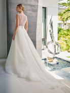 A woman wearing a white wedding dress stands indoors near a window, showing the back of the gown with a full skirt and sheer lace upper back. The setting includes a modern interior, a small pool, and a large metallic sculpture. / Hand beaded bodice made of strings of various sized pearls with beaded edge finishing. There are sheer inserts at the side and a full illusion back bodice. The skirt is made a matte organza with flat pleats to create soft volume. A soft, pleated organza belt wraps around the waist