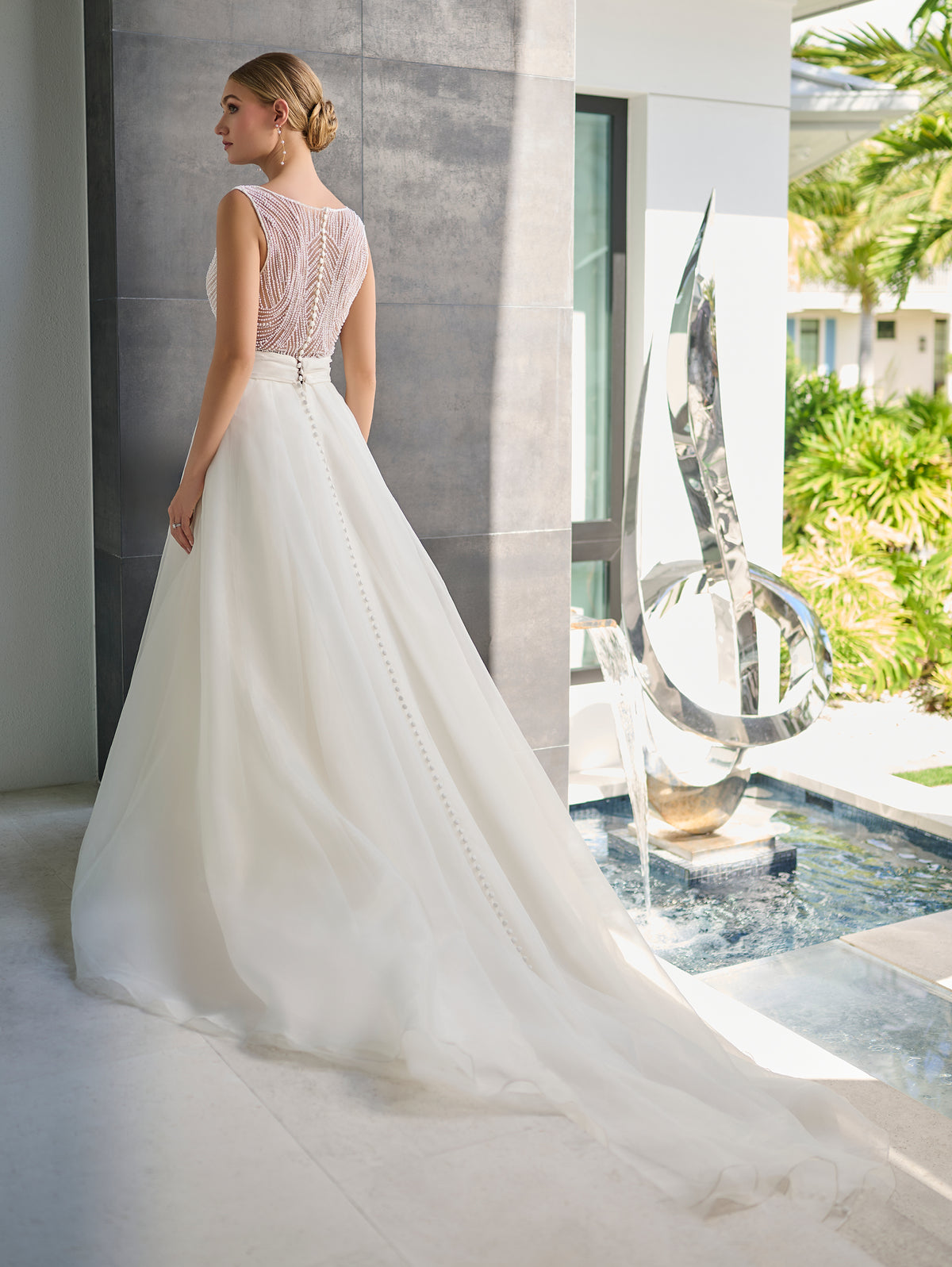 A woman wearing a white wedding dress stands indoors near a window, showing the back of the gown with a full skirt and sheer lace upper back. The setting includes a modern interior, a small pool, and a large metallic sculpture. / Hand beaded bodice made of strings of various sized pearls with beaded edge finishing. There are sheer inserts at the side and a full illusion back bodice. The skirt is made a matte organza with flat pleats to create soft volume. A soft, pleated organza belt wraps around the waist