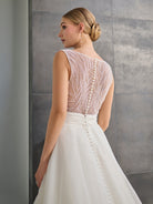 A woman modeling a sleeveless white wedding dress from the back, showing a heavily beaded and embellished back bodice with a structured waistband and flowing skirt, set against a gray tiled wall. / Hand beaded bodice made of strings of various sized pearls with beaded edge finishing. There are sheer inserts at the side and a full illusion back bodice. The skirt is made a matte organza with flat pleats to create soft volume. A soft, pleated organza belt wraps around the waist