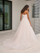 A woman wearing a strapless white wedding gown with a fitted lace bodice and a full tulle A-line skirt, photographed indoors with a large white floor and modern window blinds in the background. / This dropped waist bodice is made of a stunning 3D floral lace that has been placed over a layer of chantilly lace. The encased boning adds beautiful structure and secure fit. The dramatic skirt is made of several layers of tulles that all work effortlessly together.