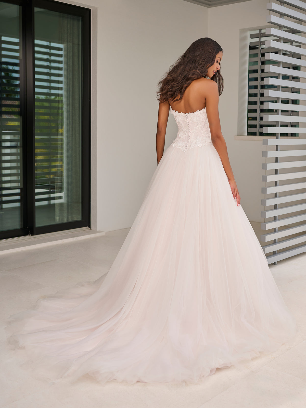 A woman wearing a strapless white wedding gown with a fitted lace bodice and a full tulle A-line skirt, photographed indoors with a large white floor and modern window blinds in the background. / This dropped waist bodice is made of a stunning 3D floral lace that has been placed over a layer of chantilly lace. The encased boning adds beautiful structure and secure fit. The dramatic skirt is made of several layers of tulles that all work effortlessly together.