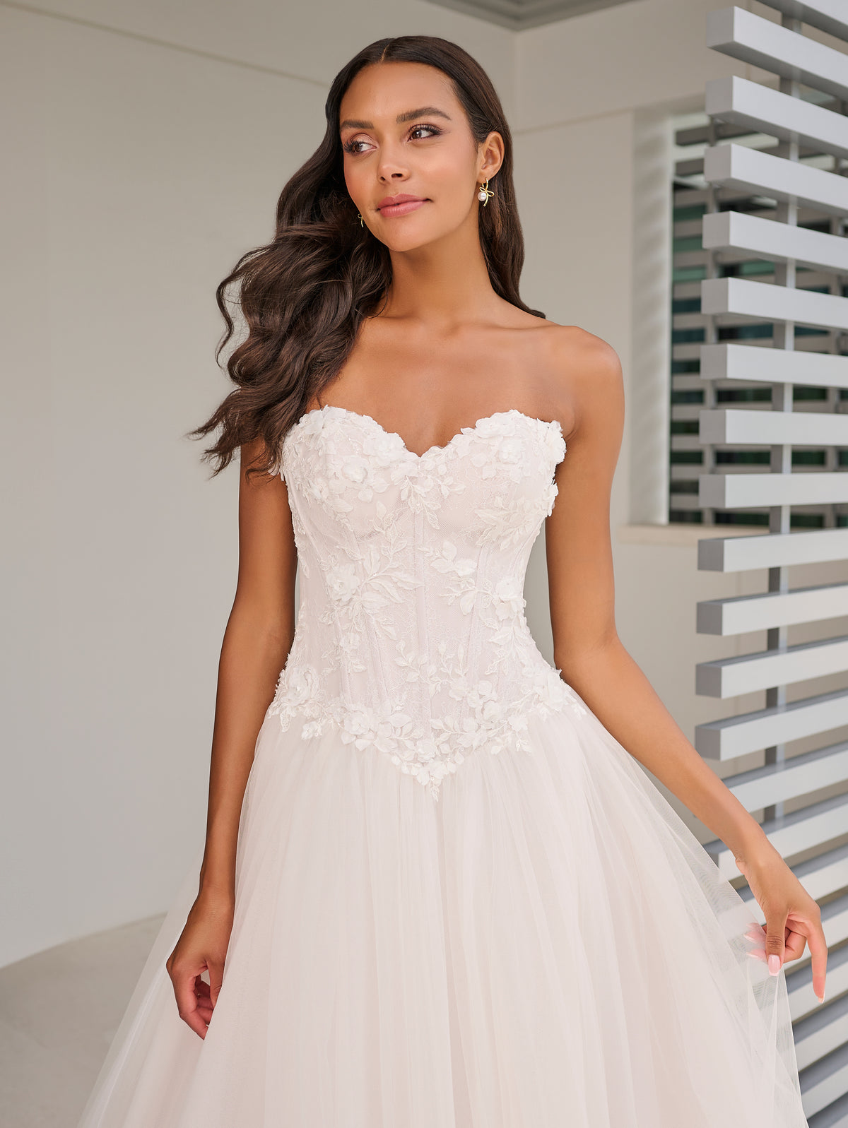 A woman modeling a strapless white wedding gown with a sweetheart neckline, floral lace applique on the bodice, and a voluminous tulle skirt in a bright indoor setting. / This dropped waist bodice is made of a stunning 3D floral lace that has been placed over a layer of chantilly lace. The encased boning adds beautiful structure and secure fit. The dramatic skirt is made of several layers of tulles that all work effortlessly together.