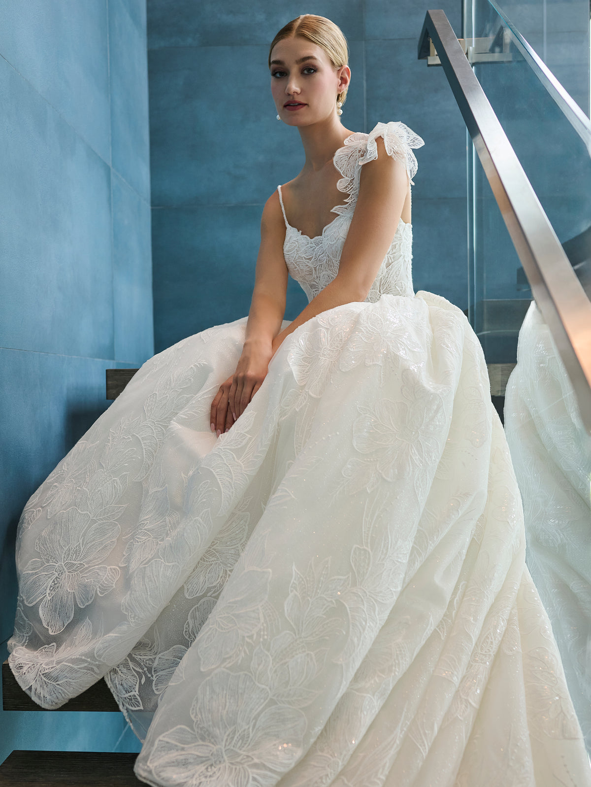 A woman wearing a voluminous white wedding gown with floral lace applique and ruffled shoulder details, seated on a staircase in a modern interior with blue walls. / This is a stunning ball gown made out of floral all over lace with glitter mesh underlayer. Semi-sheer bodice with a scooped neckline, beaded spaghetti straps and beautiful lace feature on one shoulder.
