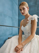 A woman wearing a white lace bridal gown with ruffled shoulder details, seated against a blue paneled wall. / This is a stunning ball gown made out of floral all over lace with glitter mesh underlayer. Semi-sheer bodice with a scooped neckline, beaded spaghetti straps and beautiful lace feature on one shoulder.