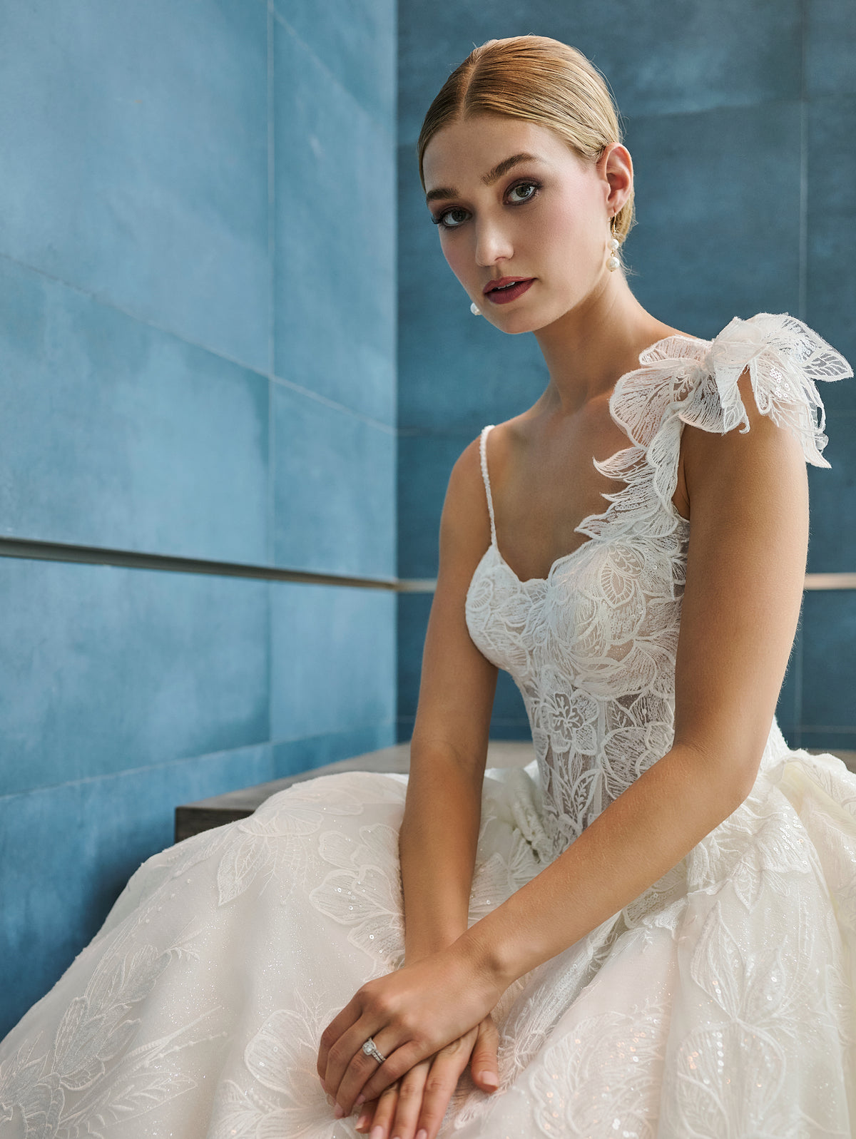 A woman wearing a white lace bridal gown with ruffled shoulder details, seated against a blue paneled wall. / This is a stunning ball gown made out of floral all over lace with glitter mesh underlayer. Semi-sheer bodice with a scooped neckline, beaded spaghetti straps and beautiful lace feature on one shoulder.