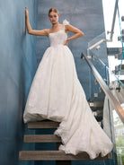 A woman wearing a white lace ball gown wedding dress stands on a staircase indoors, with one hand resting against a blue-gray wall and the other on her hip. The dress has thin straps, a fitted lace bodice, and a full layered tulle skirt with a train. / This is a stunning ball gown made out of floral all over lace with glitter mesh underlayer. Semi-sheer bodice with a scooped neckline, beaded spaghetti straps and beautiful lace feature on one shoulder.