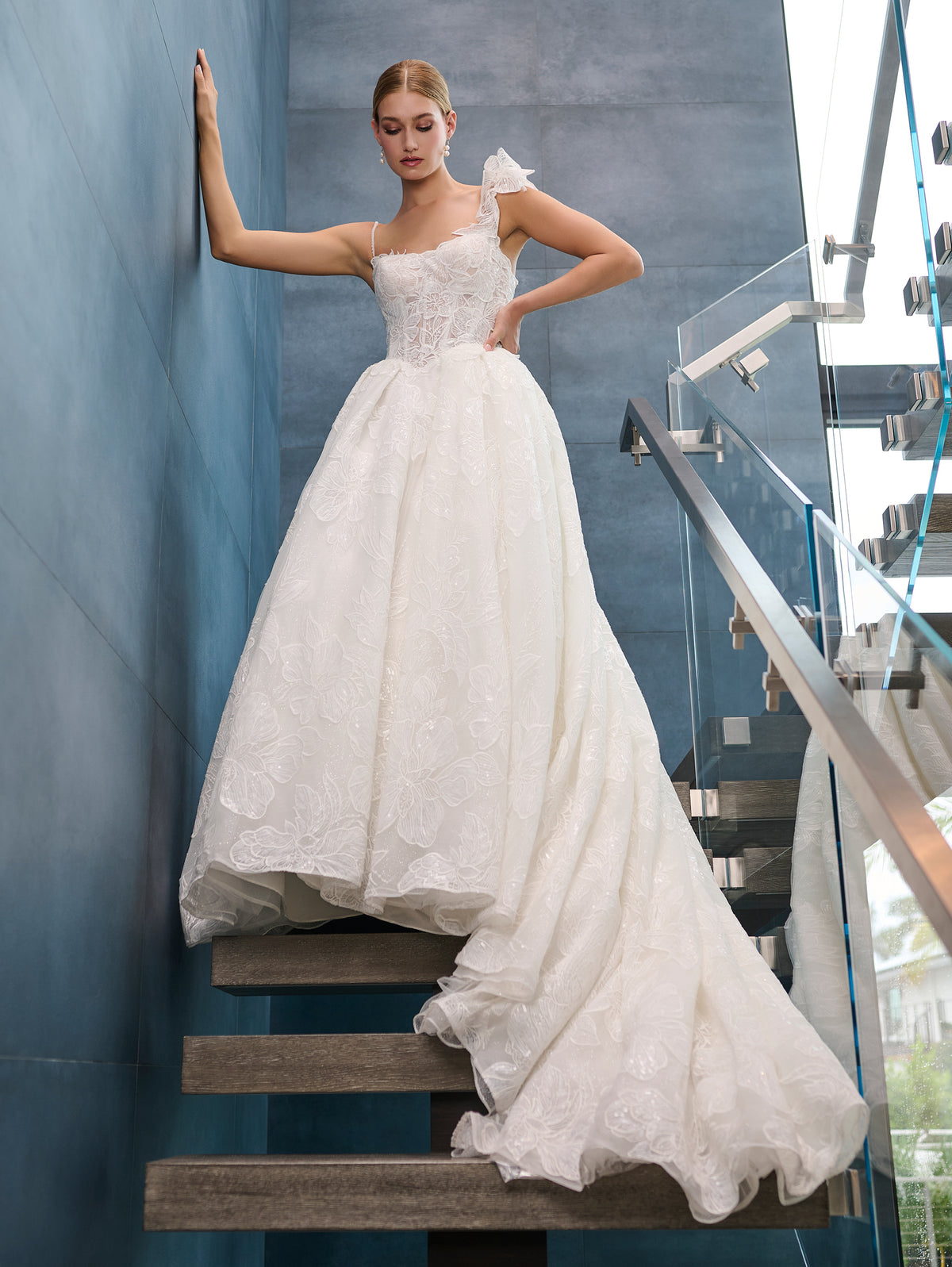 A woman wearing a white lace ball gown wedding dress stands on a staircase indoors, with one hand resting against a blue-gray wall and the other on her hip. The dress has thin straps, a fitted lace bodice, and a full layered tulle skirt with a train. / This is a stunning ball gown made out of floral all over lace with glitter mesh underlayer. Semi-sheer bodice with a scooped neckline, beaded spaghetti straps and beautiful lace feature on one shoulder.