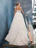 A woman wearing a white lace A-line wedding gown with thin straps and a bow detail on one shoulder, photographed from behind as she stands on a staircase indoors. / This is a stunning ball gown made out of floral all over lace with glitter mesh underlayer. Semi-sheer bodice with a scooped neckline, beaded spaghetti straps and beautiful lace feature on one shoulder.