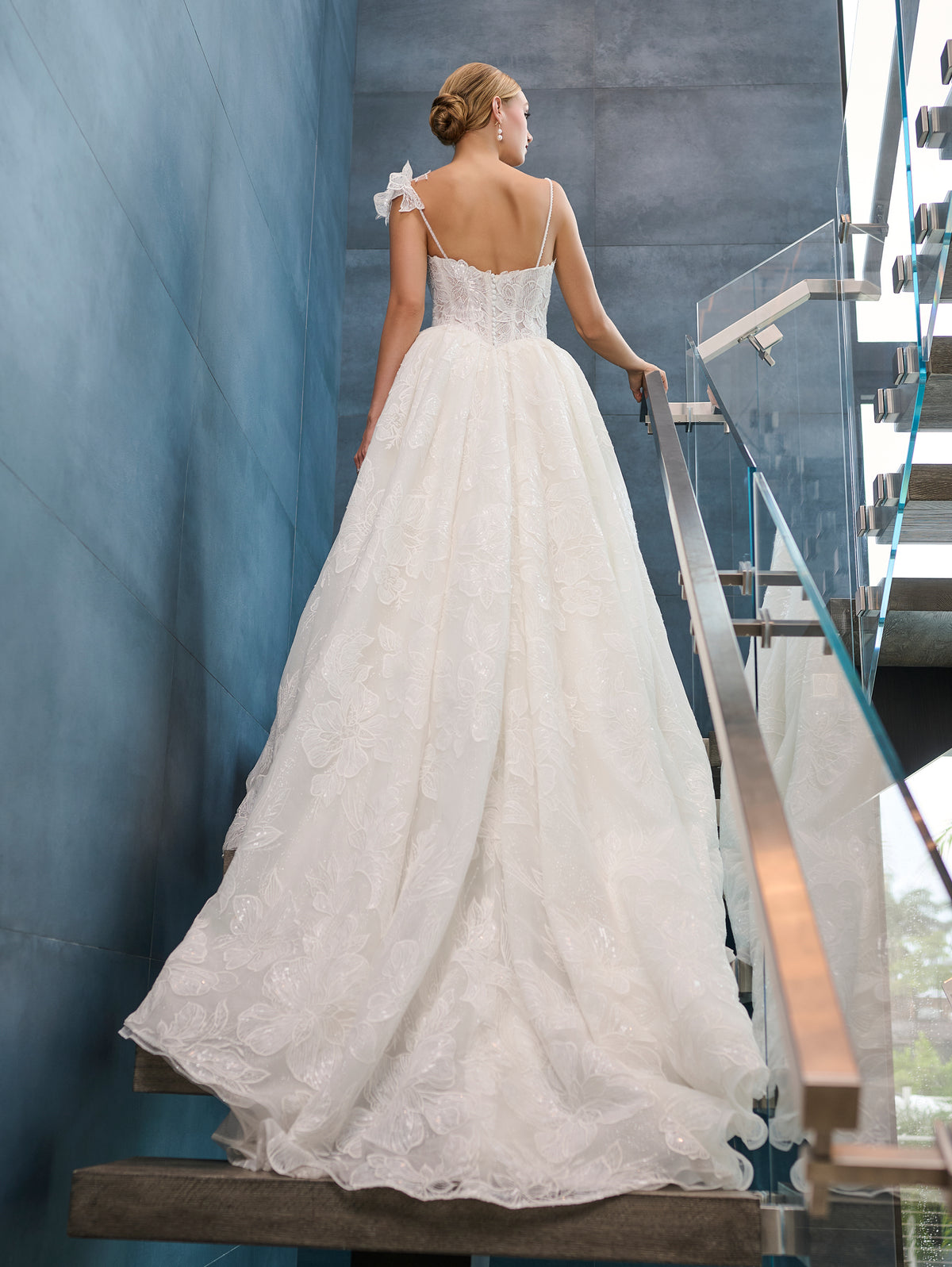 A woman wearing a white lace A-line wedding gown with thin straps and a bow detail on one shoulder, photographed from behind as she stands on a staircase indoors. / This is a stunning ball gown made out of floral all over lace with glitter mesh underlayer. Semi-sheer bodice with a scooped neckline, beaded spaghetti straps and beautiful lace feature on one shoulder.