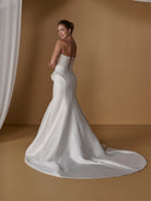 A woman modeling a strapless white satin mermaid-style wedding gown with a train and lace-up back, posed in a warm tan studio setting with draped fabric at the side. / Stretch mikado slim gown with lace and peplum detailing and lace up back.