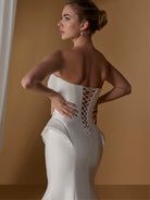 Woman wearing a white strapless gown with intricate back design against a brown background / Stretch mikado slim gown with lace and peplum detailing and lace up back.