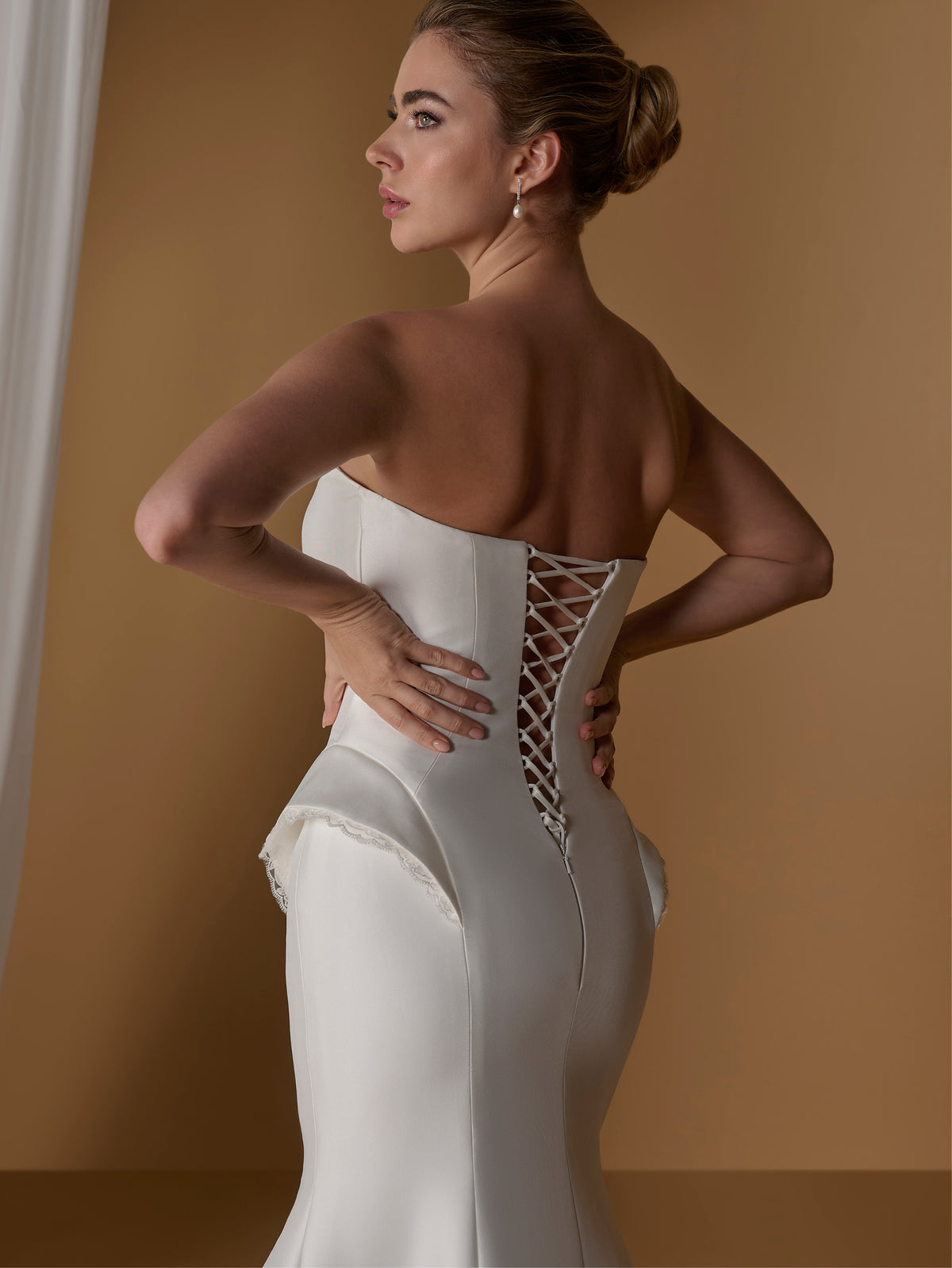 Woman wearing a white strapless gown with intricate back design against a brown background / Stretch mikado slim gown with lace and peplum detailing and lace up back.