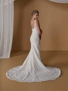 A woman modeling a fitted white lace wedding gown with a long train, photographed in a warm brown studio setting with draped white fabric on the left. / All over lace gown with square neckline.
