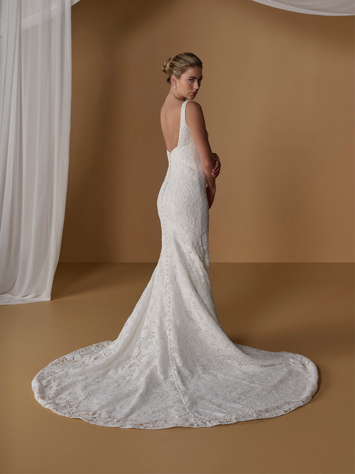 A woman modeling a fitted white lace wedding gown with a long train, photographed in a warm brown studio setting with draped white fabric on the left. / All over lace gown with square neckline.