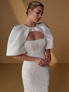 A woman wearing a white lace wedding dress with dramatic structured satin cape sleeves, posed against a warm tan studio backdrop. / All over lace gown with square neckline.