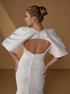 A woman shown from the back wearing a fitted ivory lace dress with dramatic draped cape sleeves and an open back, posed against a warm tan studio backdrop. / All over lace gown with square neckline.