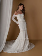 An off-the-shoulder lace wedding gown with long sheer sleeves and a flared mermaid silhouette with ruffle trim, shown on a model against a warm brown studio backdrop with a white curtain. / Slim, long sleeve lace gown with lace ruffle detail.