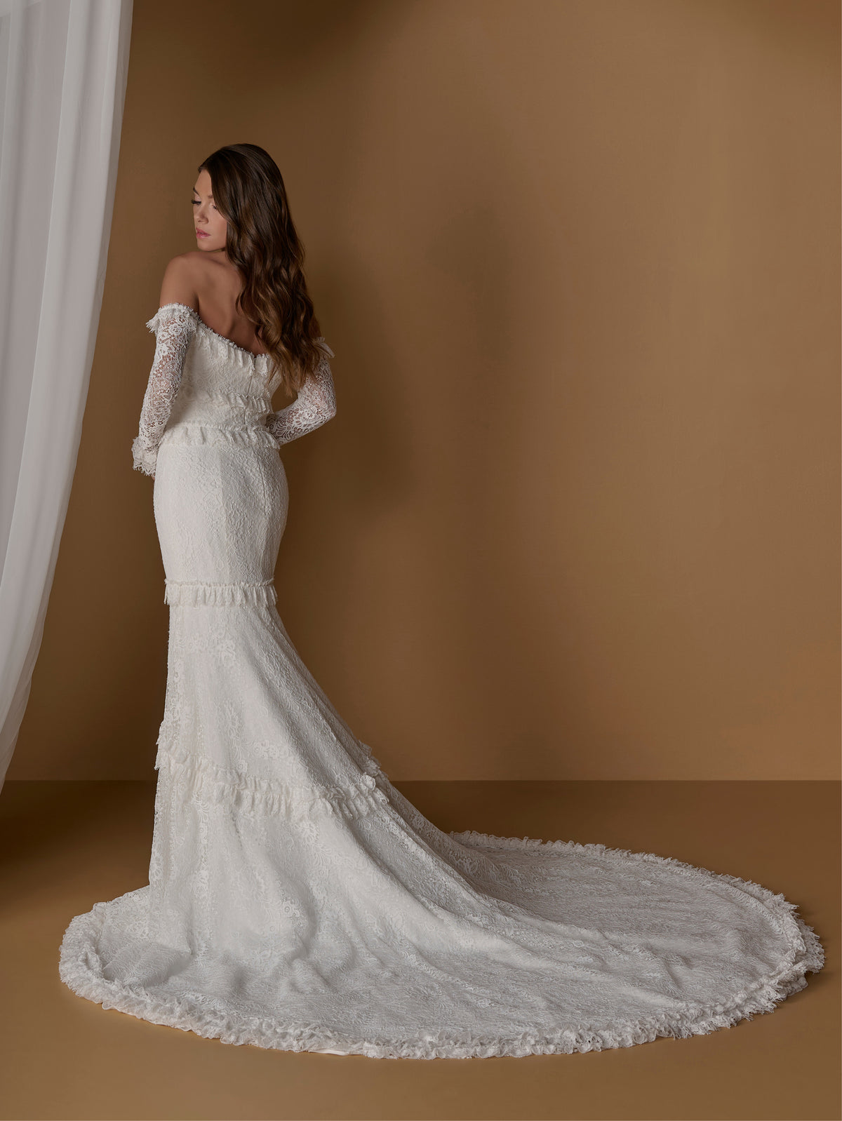 A woman wearing an off-the-shoulder white lace wedding gown with long sleeves and a dramatic train, photographed in a studio with a warm tan backdrop and white curtain on the left. / Slim, long sleeve lace gown with lace ruffle detail.