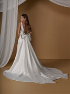 A woman modeling a white satin bridal gown with a dramatic train and large bow detail on the back, posed against a warm brown backdrop with draped white fabric. / A-line ballgown with modern dart detailing, illusion back and detachable bow.