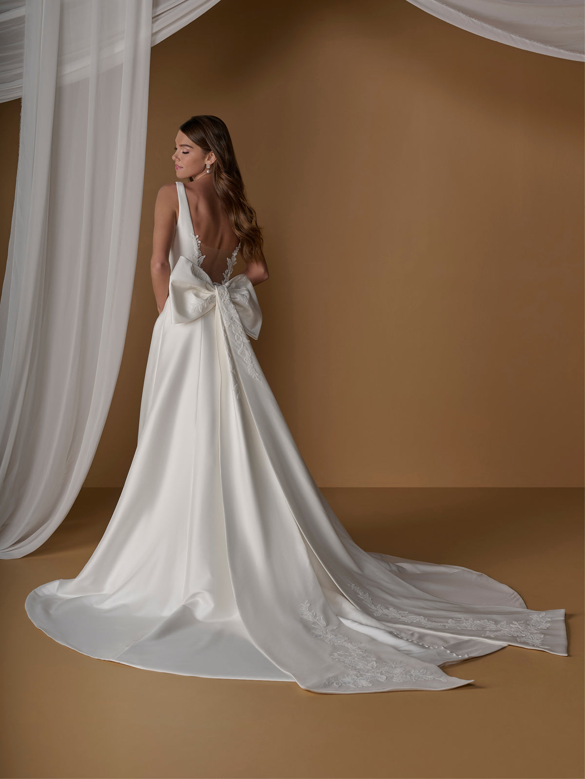 A woman modeling a white satin bridal gown with a dramatic train and large bow detail on the back, posed against a warm brown backdrop with draped white fabric. / A-line ballgown with modern dart detailing, illusion back and detachable bow.