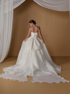 A woman wearing a strapless white wedding gown with a large bow at the back and a long flowing train, photographed in a studio with a warm tan backdrop and draped white fabric. / Satin faced organza printed ballgown with dramatic skirt and bow jacket.
