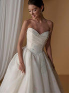 A woman modeling a strapless sweetheart ball gown with a heavily draped bodice and a full skirt featuring a subtle pastel floral print, set against a warm beige background with a white curtain. / Satin faced organza printed ballgown with dramatic skirt and bow jacket.