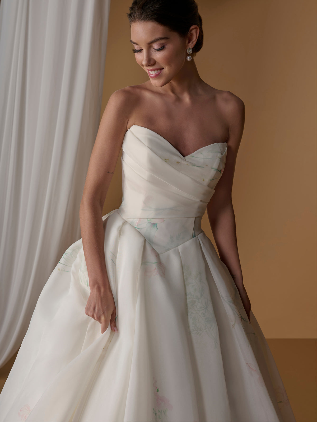 A woman modeling a strapless sweetheart ball gown with a heavily draped bodice and a full skirt featuring a subtle pastel floral print, set against a warm beige background with a white curtain. / Satin faced organza printed ballgown with dramatic skirt and bow jacket.