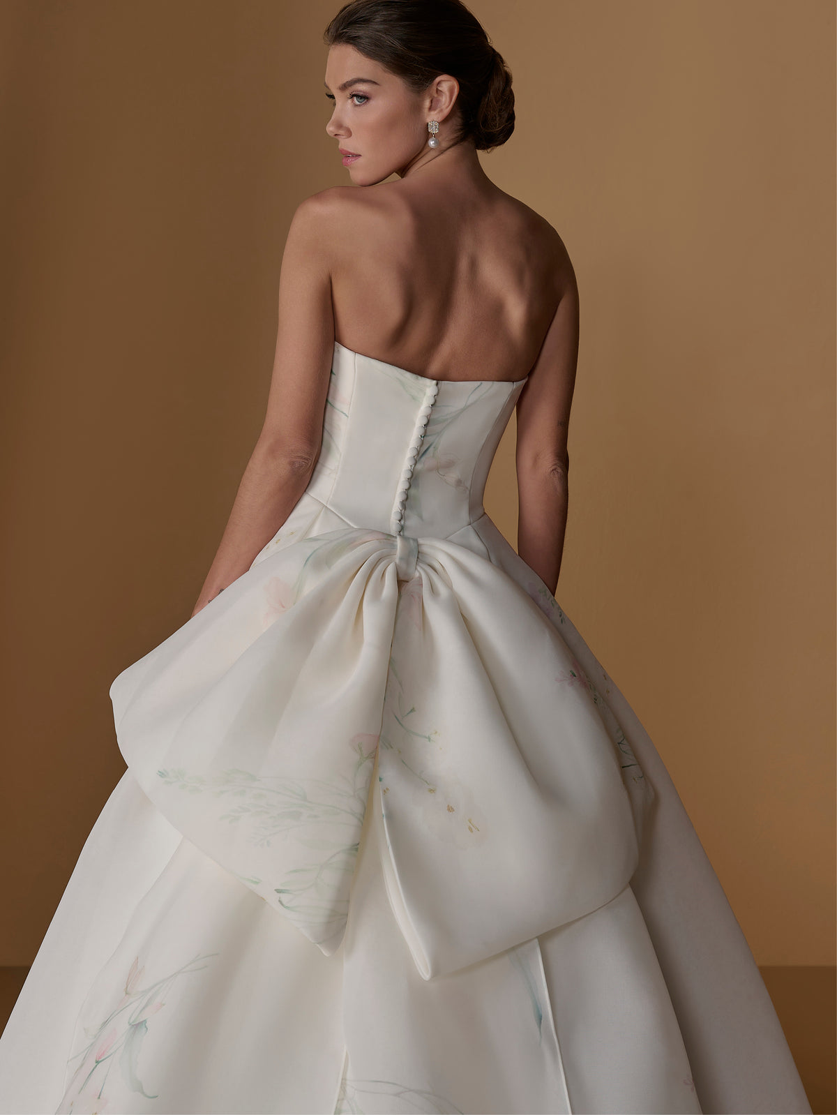 A woman modeling a strapless white ball gown from the back, featuring a structured corset bodice, a large bow at the back, and a voluminous skirt with subtle pastel floral accents, set against a warm tan studio backdrop. / Satin faced organza printed ballgown with dramatic skirt and bow jacket.