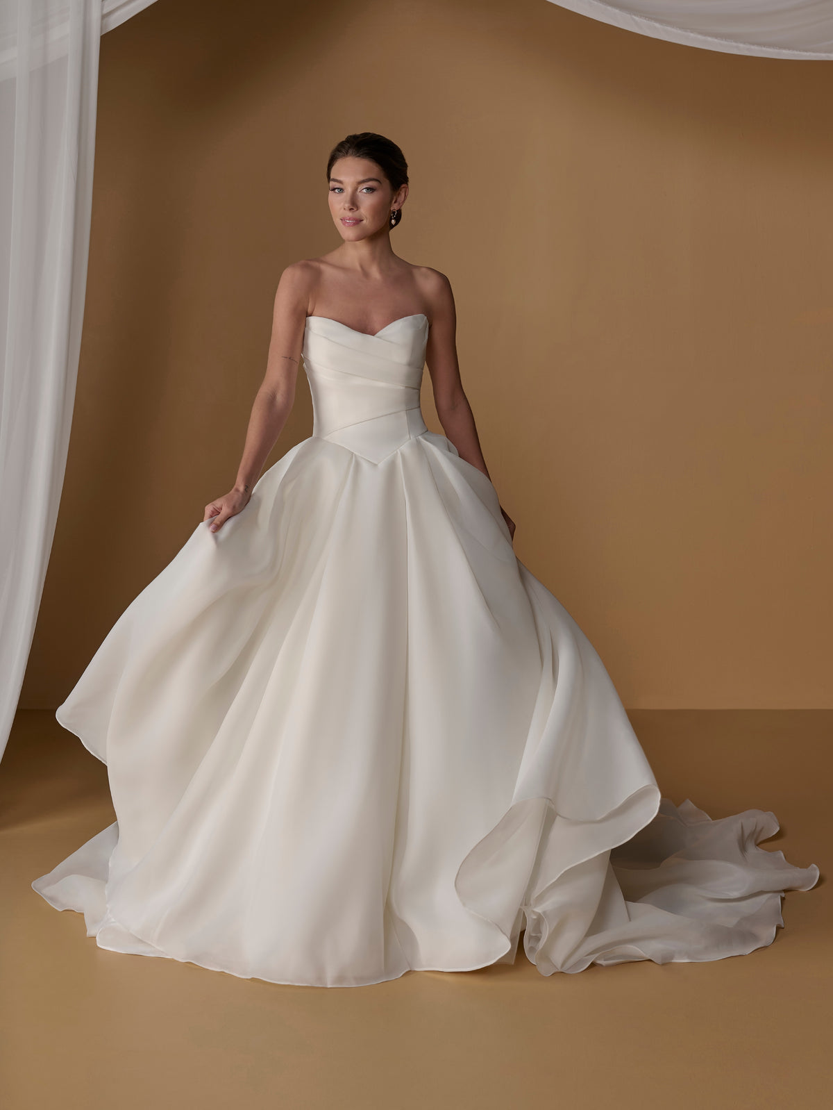 A woman modeling a strapless white ball gown wedding dress with a sweetheart neckline, structured bodice, and full layered skirt, posed against a warm tan studio backdrop with draped white fabric at the sides. / Satin faced organza ballgown with dramatic skirt and bow jacket.