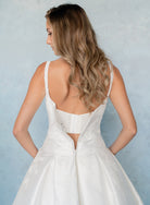 A woman wearing a white wedding dress, shown from the back against a textured light blue background. / Drop waist, mikado ballgown with pleating detail and scattered lace applique.
