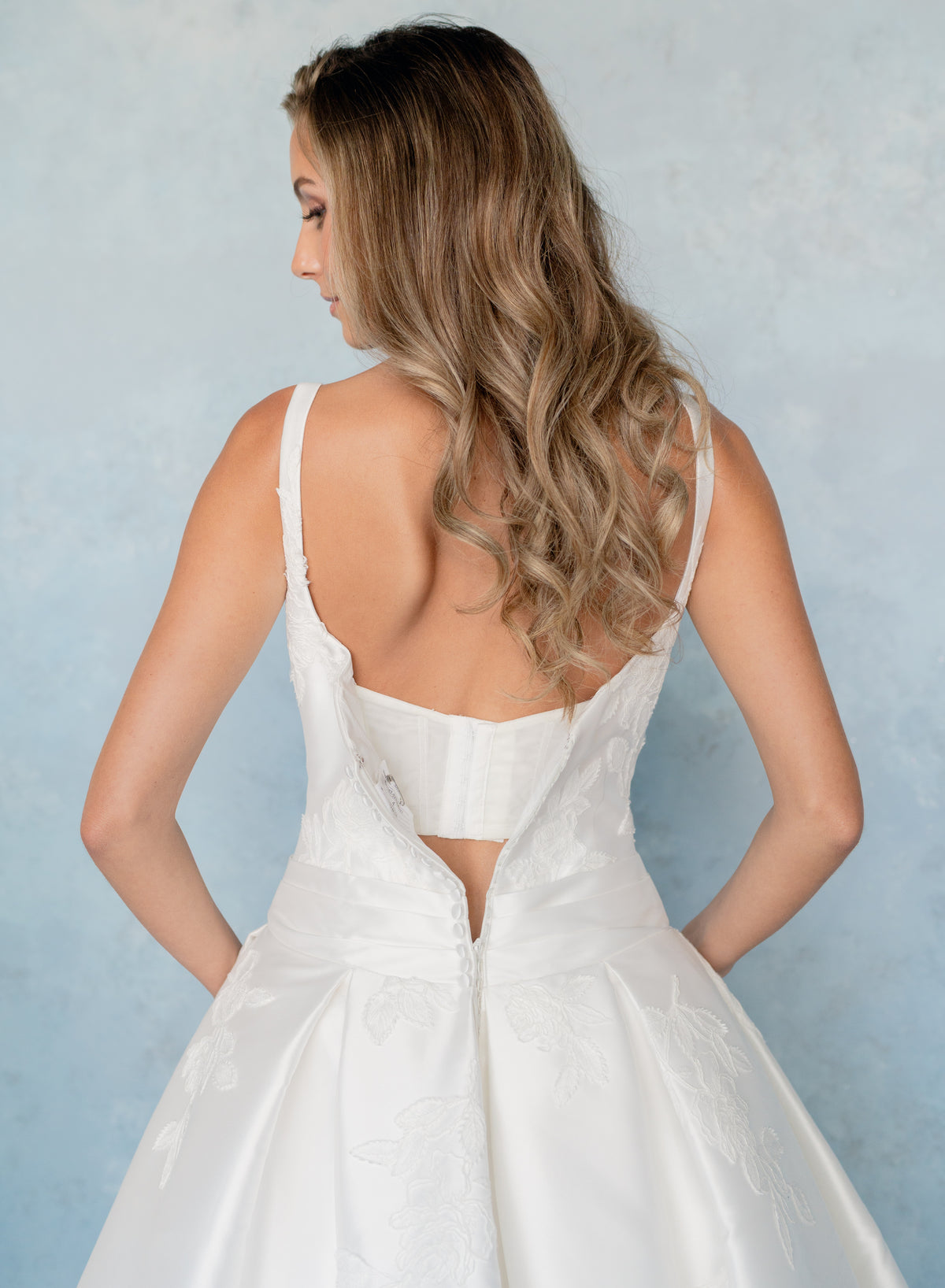 A woman wearing a white wedding dress, shown from the back against a textured light blue background. / Drop waist, mikado ballgown with pleating detail and scattered lace applique.