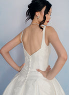 A woman wearing a white sleeveless bridal gown with a deep open back, pearl-style back buttons, and a full skirt, photographed against a soft light blue studio background. / Drop waist, mikado ballgown with pleating detail and scattered lace applique.