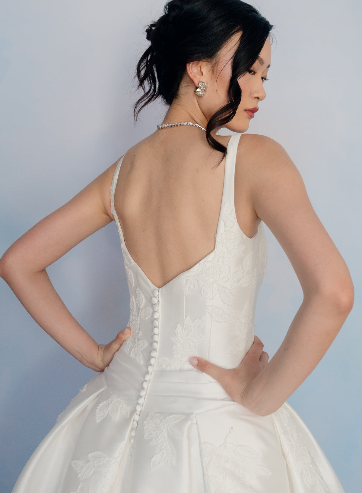 A woman wearing a white sleeveless bridal gown with a deep open back, pearl-style back buttons, and a full skirt, photographed against a soft light blue studio background. / Drop waist, mikado ballgown with pleating detail and scattered lace applique.