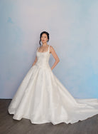 A woman modeling a white sleeveless wedding gown with a fitted bodice, wide full skirt, and long train against a soft blue studio backdrop. / Drop waist, mikado ballgown with pleating detail and scattered lace applique.