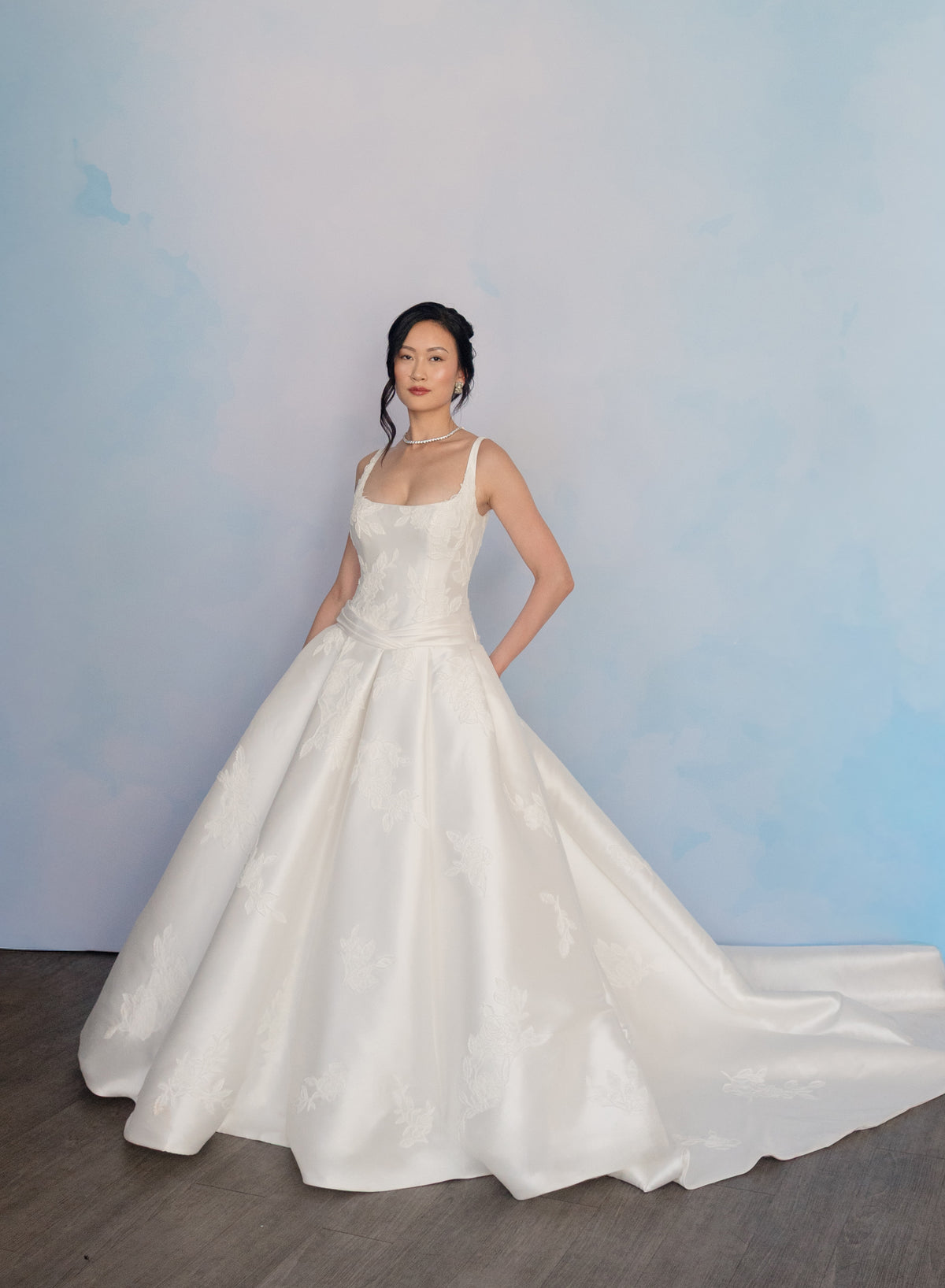 A woman modeling a white sleeveless wedding gown with a fitted bodice, wide full skirt, and long train against a soft blue studio backdrop. / Drop waist, mikado ballgown with pleating detail and scattered lace applique.