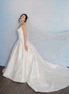 A woman wearing a white satin wedding gown with a long sheer veil, standing in a studio against a soft blue backdrop. / Lace and tulle veil.