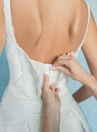 Close-up of a bride's back as she adjusts the back zipper of her white lace wedding dress against a soft light blue background. / Drop waist, mikado ballgown with pleating detail and scattered lace applique.