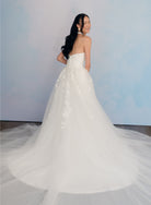 A woman wearing a strapless white wedding gown with a full tulle skirt and long train, photographed against a soft light blue studio backdrop. / Satin mermaid gown with lace appliques and detachable overskirt.