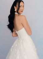 A woman wearing a strapless white wedding dress with floral lace applique and a full tulle skirt, photographed from the back against a soft light blue studio background. / Satin mermaid gown with lace appliques and detachable overskirt.