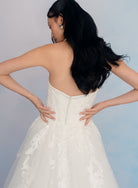 A woman wearing a strapless white wedding dress, photographed from the back against a soft light blue studio background. / Satin mermaid gown with lace appliques and detachable overskirt.