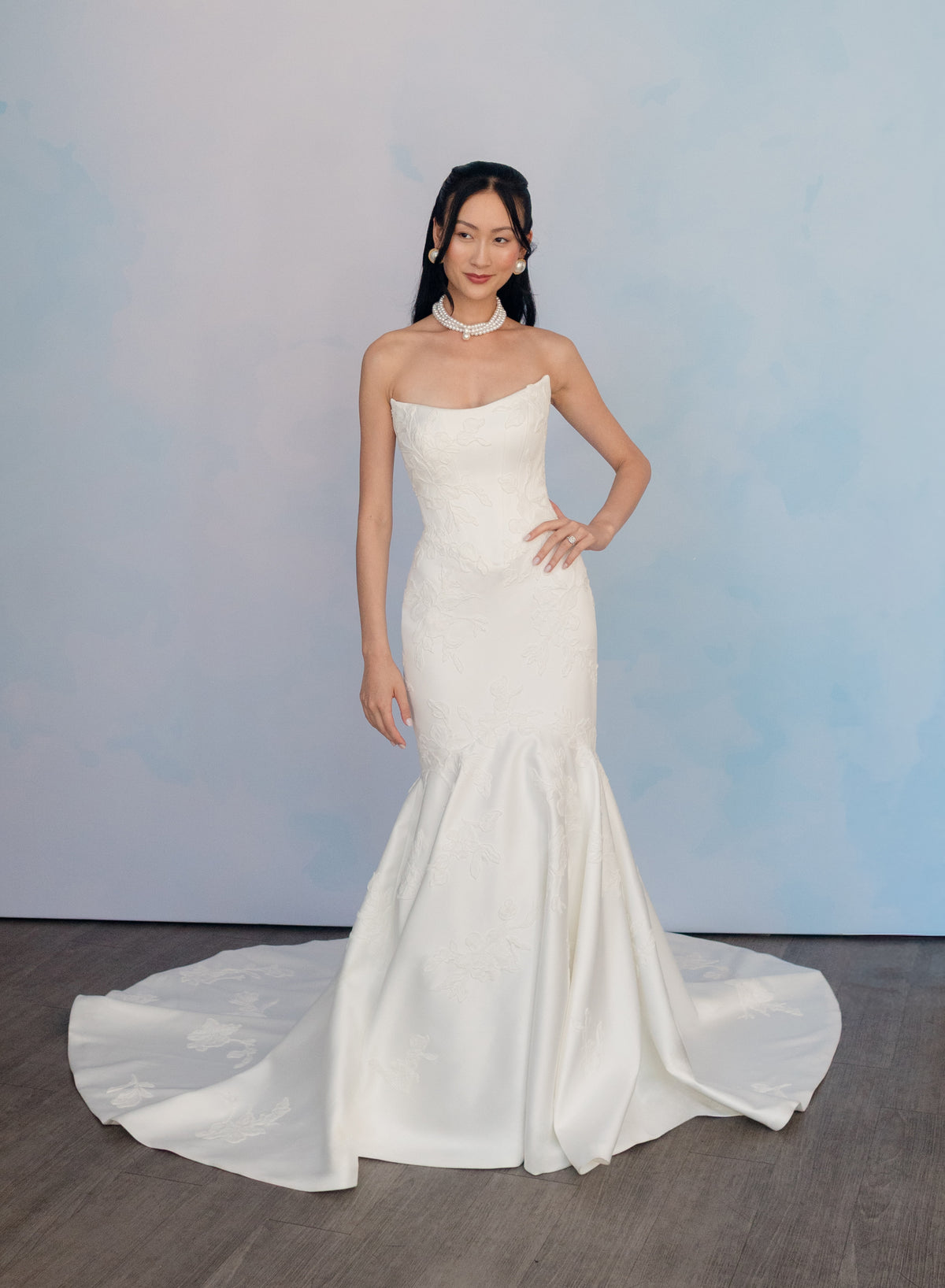 A woman modeling a strapless white mermaid-style wedding gown with a fitted bodice, flared skirt, and long train against a soft blue studio backdrop. / Double face satin, mermaid gown with placed Chantilly lace applique.