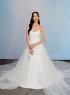 A woman modeling a strapless white wedding gown with a fitted bodice and a voluminous tulle skirt, standing against a soft blue studio backdrop. / Satin mermaid gown with lace appliques and detachable overskirt.