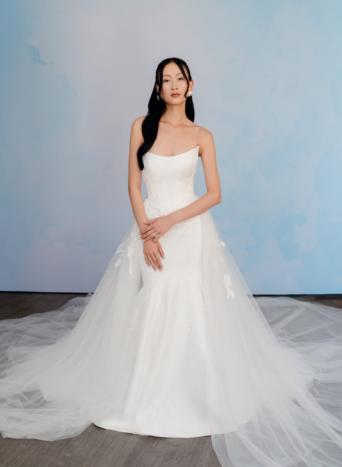 A woman modeling a strapless white wedding gown with a fitted bodice and a voluminous tulle skirt, standing against a soft blue studio backdrop. / Satin mermaid gown with lace appliques and detachable overskirt.