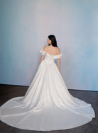 A woman wearing a voluminous off-the-shoulder white wedding gown with a long train, photographed from the back against a soft blue wall and dark wood floor. / Off the shoulder sculpted neckline gown with basque waist and full, gathered skirt.