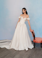A woman models an off-the-shoulder white A-line wedding gown with a sweetheart neckline, puffed sleeves, and a full skirt with a train, standing in a studio setting next to a pink chair against a light blue textured backdrop. / Off the shoulder sculpted neckline gown with basque waist and full, gathered skirt.