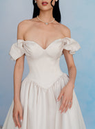 An off-the-shoulder sweetheart dress in a warm ivory or cream color, featuring a structured corset-style bodice and a flowing skirt, shown on a model against a soft blue background. / Off the shoulder sculpted neckline gown with basque waist and full, gathered skirt.